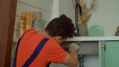 Koruyucu iş kıyafetleri içindeki Afro-Amerikan tesisatçı portresi ve mutfak musluğunu tamir eden eldivenler çelik ayarlanabilir anahtarı gösteriyor ve içeride tesisat hizmeti verirken gülümsüyor..