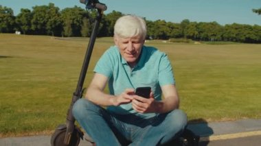 Elektrikli scooter platformunda oturan mutlu ve çekici bir adamın portresi, cep telefonu kullanarak sosyal medya içeriğini tararken parkta aktif yaşam tarzının ve yaz doğasının tadını çıkarıyor..