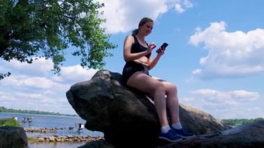 Nehir kenarındaki kayalıklarda oturan, internet üzerinden mesajlaşan, akıllı telefon kullanan ve yoğun spor eğitiminden sonra yaz doğasında dinlenen çekici atletik kadın..