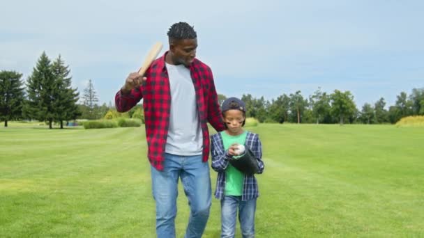 拿着棒球棒的快乐英俊的非洲父亲抱着可爱的 快乐的 年轻的非洲裔美国男孩 戴着球和棒球手套 沿着绿地散步 在棒球赛前聊天 — 图库视频影像