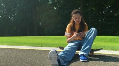 Güzel genç kaykaycı kız kaykaylı gündelik giysiler içinde kaykayın üzerinde oturuyor, internette mesajlaşıyor ve akıllı telefondan sosyal medya içeriğini tarıyor yaz parkında kaykay ve boş vakitlerin tadını çıkarıyor..