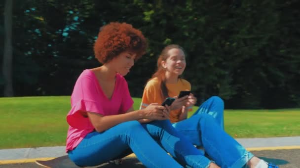 ハッピーかわいいマルチエスニック十代の娘とスケートボードに座っている魅力的な黒人の母親 携帯電話でネットワーキングし スケートボード中にレジャーを楽しんでいる間 オンラインコンテンツについて話し合う — ストック動画