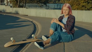 Keyifsiz, çekici, pembe saçlı kadın kaykaycı. Yaralı dizini tutuyor ve masaj yapıyor. Gün doğumunda şehir sokağında kaykay yaparken düşüşünden sonra acı ve rahatsızlık duyuyor..