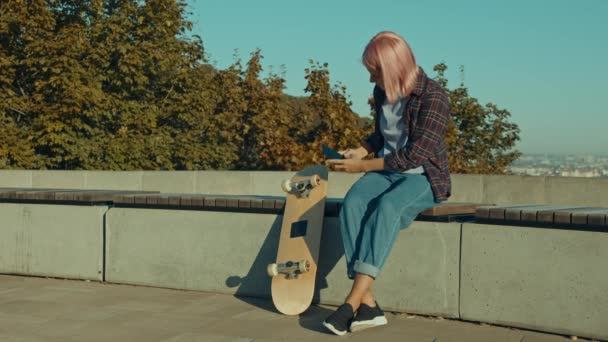 トレンドクールかわいいピンク髪の女性スケートボーダーベンチに座って 夜明けに都市の設定でスケートボードをしながら 携帯電話でスケートボードホイールの写真を撮り オンラインでスペアパーツをブラウジング — ストック動画