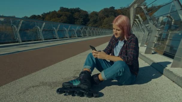 ローラースケートのポジティブかわいいピンク髪の女性 歩行者ブリッジに座って ネットワーキングとブラウジング ソーシャルメディアコンテンツ 携帯電話を使用してオンライン デイブレイクでローラーブレード — ストック動画