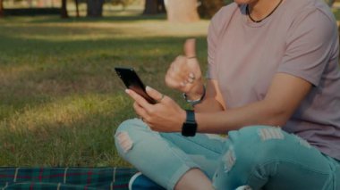 Orta yaşlı işitme engelli bir kadının cep telefonuyla video konferans aracılığıyla işaret diliyle konuşması, ekose üzerinde oturması ve halk parkında eğlenmesi.