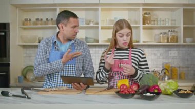Çekici işitme engelli aile önlükleriyle lezzetli yemekler pişirmeye hazırlanıyor, dijital tabletteki gerekli yemek malzemelerini kontrol ediyor ve evcil mutfakta işaret diliyle konuşuyor..