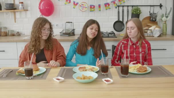 お祝いのテーブルに座っている魅力的な十分に授与された10代の女性の友人 過度のおいしいバーガー ジャガイモやナゲット 自宅で誕生日を祝う間お祝いのディナーの満足と喜びを示す — ストック動画