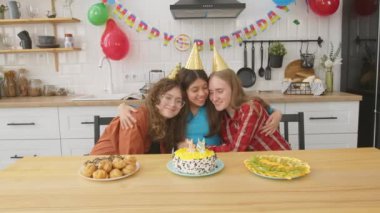 Parti şapkalı neşeli çekici genç kız neşeli genç bayan arkadaşları kucaklıyor, güçlü dostluk, birliktelik ve mutluluk ifade ederken festival masasında doğum günü partisini kutluyor..