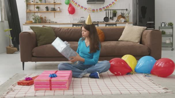 好奇心旺盛な幸せな愛らしい思春期の誕生日少女は床に座って帽子をかぶり 屋内で誕生日パーティーの後にレジャーを楽しんでしながらギフトボックスに提示しようとします — ストック動画