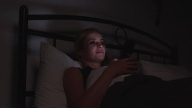 Gecelik giymiş rahat, olgun bir kadının portresi yatakta uzanıyor, sosyal medya içeriğini internette tarıyor geceleri karanlık yatak odasında dinlenirken cep telefonu kullanıyor..