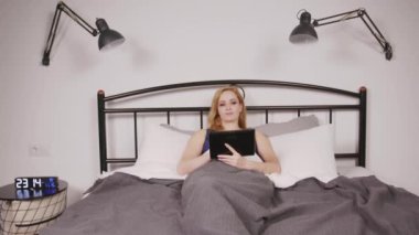 Yatmadan önce yatak odasında dinlenirken dijital tablet kullanarak sosyal medya içeriğini tarayan yatağında yatağında oturan orta yaşlı, pozitif çekici bir kadın..