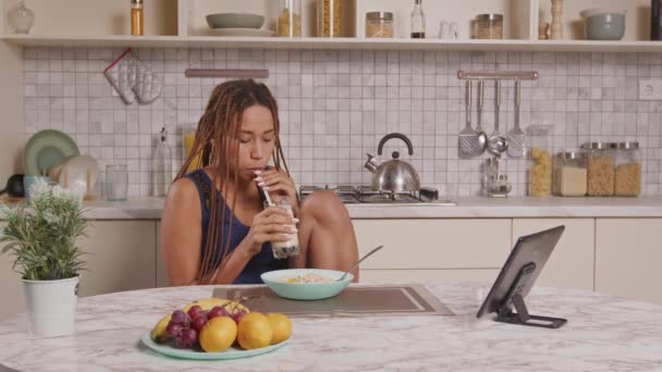 身穿睡衣的积极的黑人女性 一边吃着意大利早餐 一边吃着新鲜的水果片 一边喝着冰沙 一边上网观看视频内容 同时在家里的厨房里消闲 — 图库视频影像