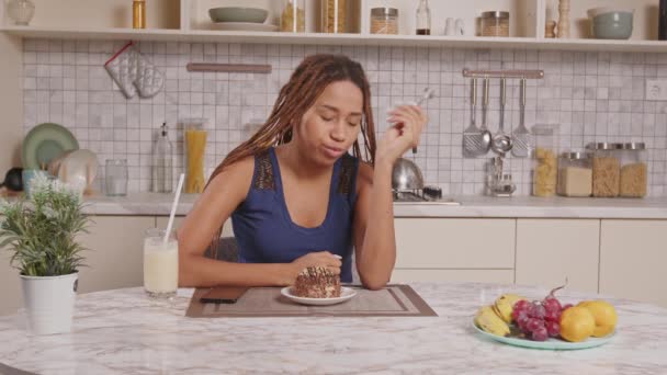 身穿时髦辫子睡衣的迷人的非洲裔美国女人坐在餐桌边 一边吃着美味的巧克力 一边在自家厨房里放松一下 — 图库视频影像