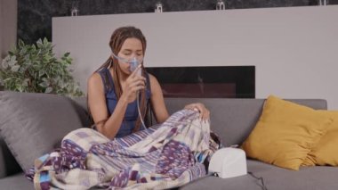 Örgü örmüş, ultrasonik dalga nebulizatörü kullanarak ilaç tedavisi uygulayan, astım ilacı soluyan, evinde kanepede otururken hasta ve sevimli Afro-Amerikan kadın..