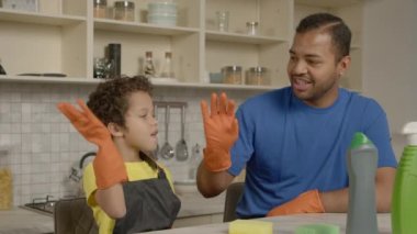 Pozitif okul çağında siyah çocuk ve koruyucu eldivenli neşeli baba bahar temizliğinden önce beş jest yapıyor, ev mutfağında hazırlık ve beraberlik gösteriyor..