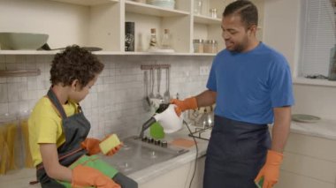 Önlüklü yakışıklı Afrikalı Amerikalı bir baba koruyucu eldivenli şirin bir çocuk ev işi yapıyor kirli gaz ocağını yıkıyor mutfakta buhar temizleyici ve deterjan kullanıyor..