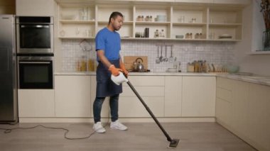 Önlüklü, koruyucu eldivenli, ev temizliği yapan, temizlik yapan, mutfak zeminini temizleyen, buharlı temizleyici kullanan yakışıklı bir adam..