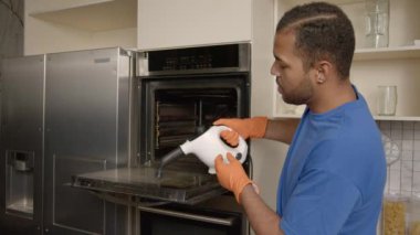 Koruyucu eldivenli ve önlüklü çekici bir Afrikalı Amerikalı ev işi yapıyor, evdeki mutfakta buhar temizleyici ve deterjan kullanarak yağdan ve yağdan kirli mutfak fırını yıkıyor..
