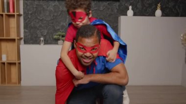 Süper kahraman maskeli ve pelerinli şirin okul çağındaki oğlunun portresi çömelmiş pozisyonlarda oturan mutlu babalar, neşeli gülümsemelerle, kapalı alanda neşeli ruh hali ve neşe ifade ederek.