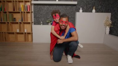 Pozitif yakışıklı siyah baba çömelme pozisyonunda ve sevimli prematüre oğlu süper kahraman maskeleri ve pelerinler takıyor, içeride eğleniyor ve neşeli gülücükler atıyor..