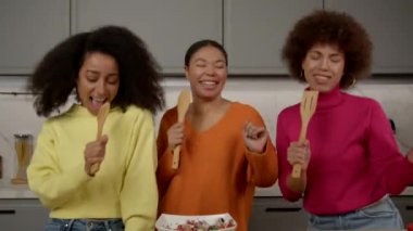 Neşeli Afro-Amerikan kadınlarının portresi spatulalarla dans edip şarkı söylüyor, ev mutfağında yemek pişirirken kaygısız ruh halini ve mutluluğu ifade ediyor..