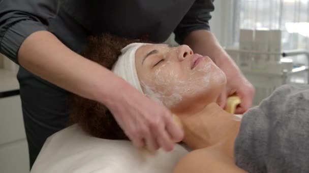 美しいアフリカ系アメリカ人女性が顔の皮膚治療を受け スパサロンで熟練した化粧師によるスポンジによる若返りの皮膚表面剥離マスク — ストック動画