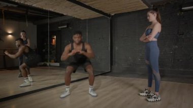 Motive olmuş aktif spor formundaki siyah adam, kadın fitness eğitmeninin rehberliği altında eğilip, vücut kaslarını güçlendiriyor ve sağlık kulübünde istikrar kazanıyor..