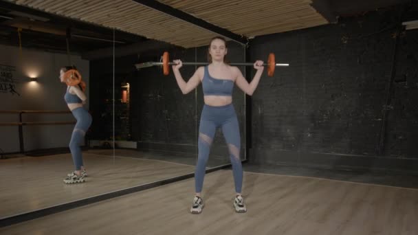 体重トレーニングを実践するスポーツウェアのモチベーションのスリムな魅力的なフィット感の女性を決定し バーベルスクワット運動を行い より低い体を強化し グラートと四肢をターゲットにします ヘルスクラブで — ストック動画
