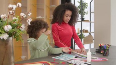 Çekici siyah anne ve sevimli okul çağı kıvırcık Afrikalı Amerikalı kız yaratıcı ve sanatsal çocuk becerileri geliştiriyor, renkli kağıt şeritleri seçerek el işi tasarımı yapıyor.