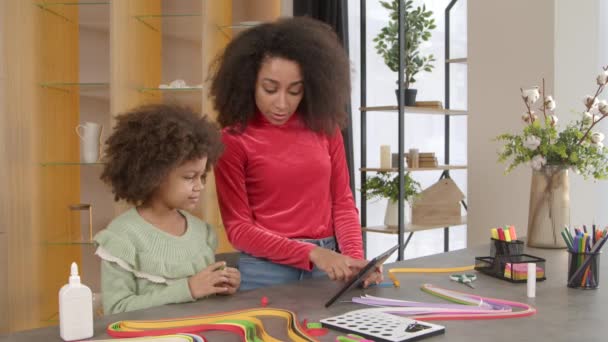 有爱心的非洲裔美籍母亲和可爱的小卷曲女儿进行交流 一边做手工装饰一边看数码平板电脑补丁的网上视频教学 — 图库视频影像
