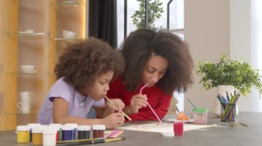 Neşeli kıvırcık saçlı, kıvırcık saçlı Amerikalı anne ve sevimli ilkokul kızı birlikte resim yapıyorlar. İçerideki sanat eserlerinde ilham ve canlı hayal gücünü ifade ediyorlar..