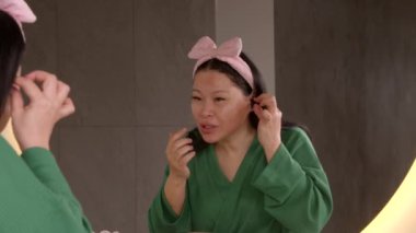 Bornozlu neşeli, orta yaşlı Asyalı kadın duştan sonra aynada görünüşünü kontrol ediyor, akıllı telefondan arkadaşlarıyla tuvalette cep telefonu kullanmadan konuşuyor. .
