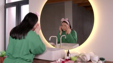 Çekici, orta yaşlı bir Asyalı kadın sabahlıkla aynadaki yansımasına bakıyor, yüz kırışıklıklarını ve cildini kontrol ediyor, banyodaki yaşlanma süreciyle ilgili endişe duyuyor..