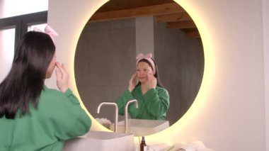 Çekici, orta yaşlı, elbise giymiş, aynada yansımaya bakan Asyalı kadın, yüz masajı ve canlandırma egzersizi yapıyor, yüz hatlarını şekillendiriyor ve cildini rahatlatıyor.