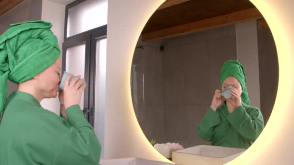 リラックスしたポジティブな魅力的なアジアの女性のバスローブとタオルは 鏡で彼女の外観を楽しんで頭の周りに包まれ コーヒーを飲んで バスルームで顔の霧スプレーを加える — ストック動画