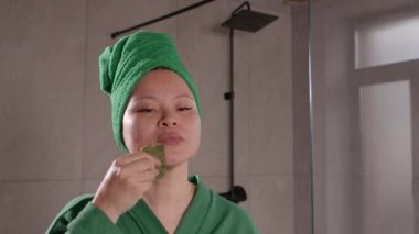 Kafasına elbise ve havlu sarılmış orta yaşlı güzel bir Asyalı kadının portresi, spa gua sha tedavisi ve yüz gerdirme işlemi yapıyor. Banyoda yeşim yüz masajı aleti kullanarak..