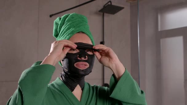 魅力的な成熟したアジアの女性の肖像画 鏡を見て スキンケアのためのスパ手順を仕上げ 黒水和顔面シートマスクを取り外し バスルームで外観を楽しむ — ストック動画