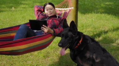 Çekici orta yaşlı Asyalı kadın hamakta uzanıyor. İtaatkar, sevimli siyah çoban köpeğiyle birlikte dışarıda eğlenirken tablet bilgisayarından online içerik izliyor..