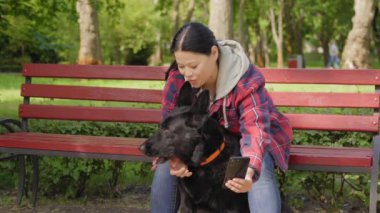 Sevecen, orta yaşlı Asyalı kadın bankta oturan sevimli siyah kadın çoban, cep telefonuyla sarılıyor ve selfie çekiyor, köpekle parkta dinlenirken..