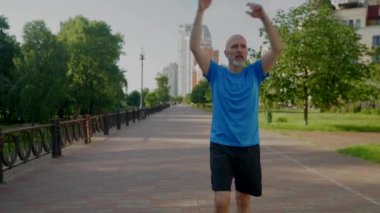 Aktif motivasyonlu sportif formda orta yaşlı bir adam ısınıyor ve kaslarını kasıyor. Sabah koşusuna hazırlanıyor. Şehir sokağında güneş ışığının altında spor antrenmanı yapıyor..