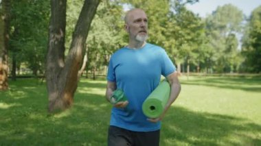 Pozitif yakışıklı, orta yaşlı, yoga minderli ve elinde su şişesi olan orta yaşlı bir adam yeşil çimlerde yürüyor, halka açık parkta yoğun bir spor salonuna gidiyor..