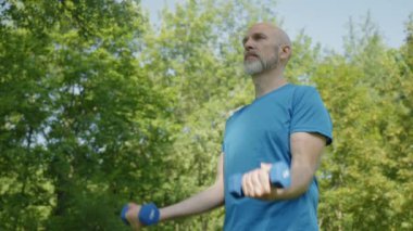 Aktif motivasyonlu çekici sporun portresi olgun bir adama uyuyor. Dambıl kıvırıyor, kolunun üst kısmını güçlendiriyor. Açık havada çalışırken kararlılık ve kendine güvenini gösteriyor..