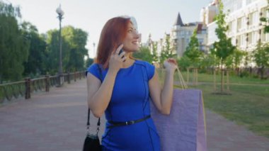 Pozitif, rahat, orta yaşlı, çekici, şık elbiseli, alışveriş torbaları taşıyan, arkadaşlarıyla cep telefonunda sohbet eden ve sokakta yürürken alışveriş günlerini paylaşan bir kadın..