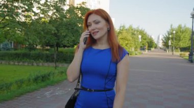 Çekici kızıl saçlı orta yaşlı kadın portresi iş kıyafetleri içinde şehir caddesinde yürüyor, cep telefonuyla arkadaşlarıyla iletişim kuruyor, pozitifliğini ve neşeli ruh halini ifade ediyor..