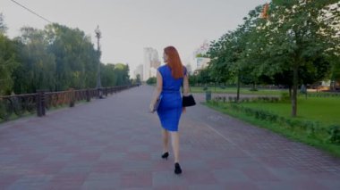 Kendine güvenen, zarif, kızıl saçlı, yüksek topuklu ayakkabılı, şık bir elbise giyen, elinde belgeler ve dijital tablet olan olgun iş kadını işe yaya caddesinde yürüyor..