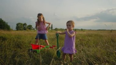 Pozitif, sevimli ilkokul kızı ve el arabasıyla gezen küçük kız kardeşi gün batımında kırsal kesimdeki yeşil alanda evcil hayvan için yeşil ot topluyor..