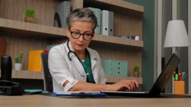 Üniformalı, üst düzey kadın pratisyen hekimin portresi masada oturuyor, bilgisayarla iletişim kuruyor ve tıp bürosunda çalışırken sağlık kontrollerini analiz ediyor..