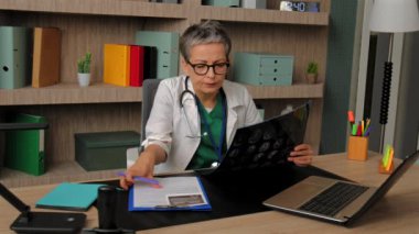 Üniformalı yaşlı bir kadın doktor ara veriyor, gözlüğünü çıkarıyor, yorgun gözlerini ovalıyor ve hasta bir hastanın muayenehane sonuçlarını analiz ediyor..