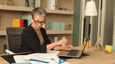 Gözlüklü, odaklanmış orta yaşlı kadın iç mimar mimari plan yapıyor, profesyonel taslak araçları kullanarak plan çiziyor ve ofiste çalışırken dizüstü bilgisayarla ağ kuruyor..